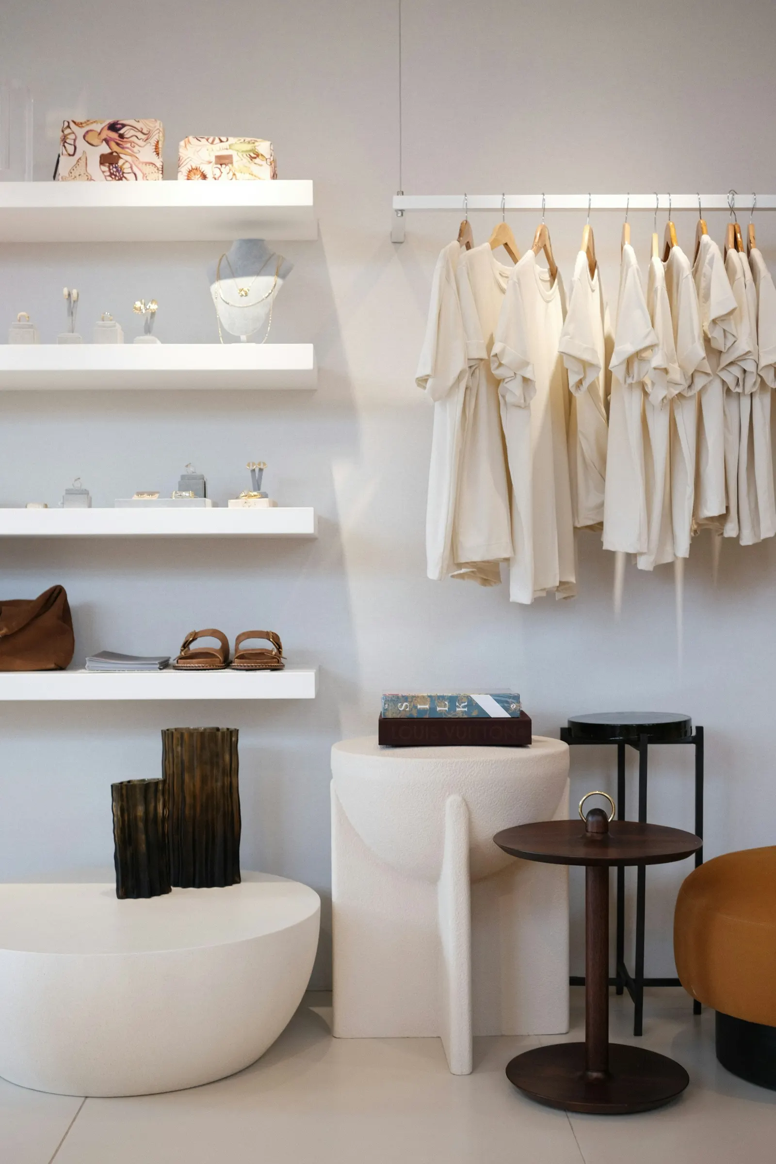 A minimal boutique interior with clothing on a rail, shoes and accessories on white shelves, and sculptural stools
