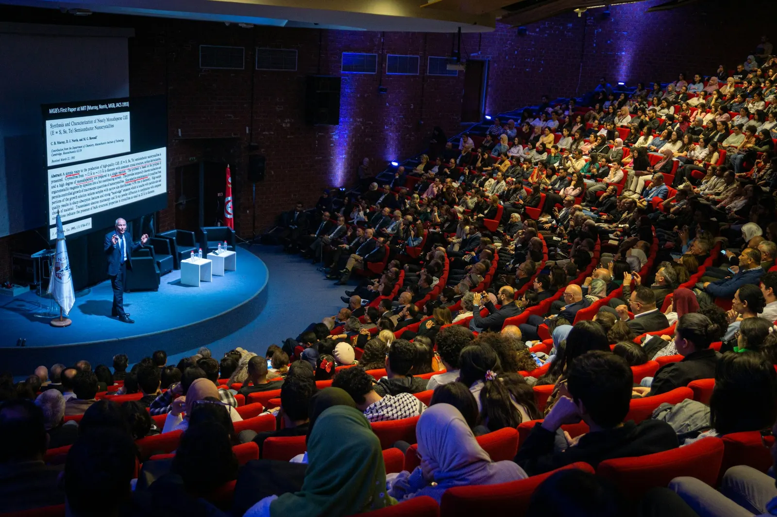 A speaker presenting on a lit stage to a full auditorium
