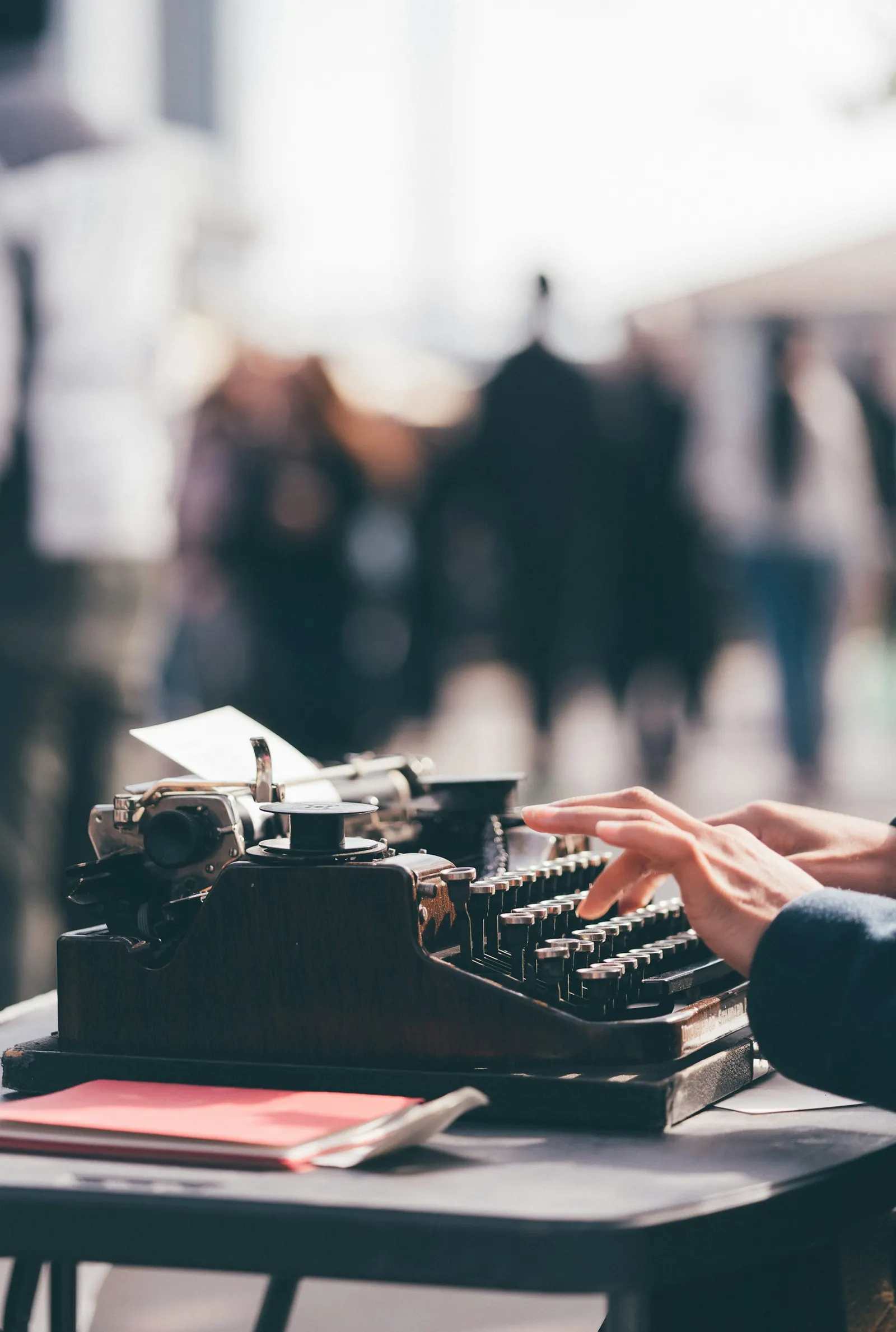 A person typing on a vintage black typewriter at an outdoor table, with a busy street blurred in the background