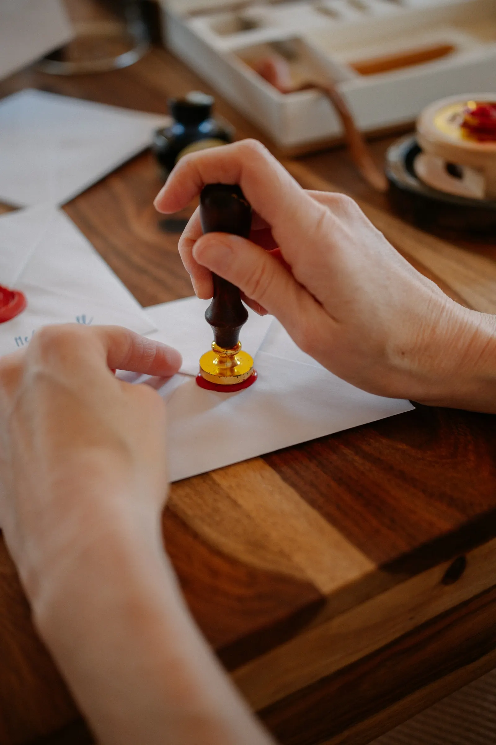 Hands pressing a wax seal stamp onto a white envelope on a wooden desk