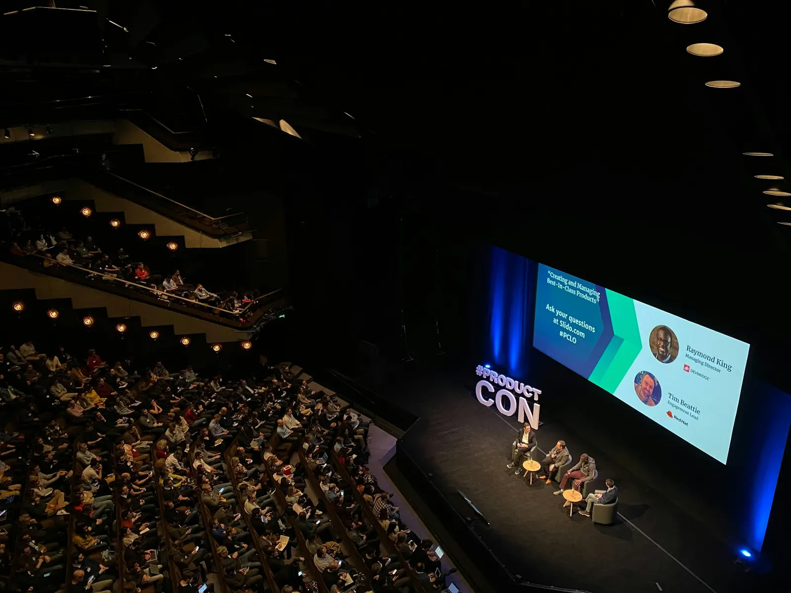 A panel discussion on a large auditorium stage, with speaker names and titles projected on the screen behind the panelists