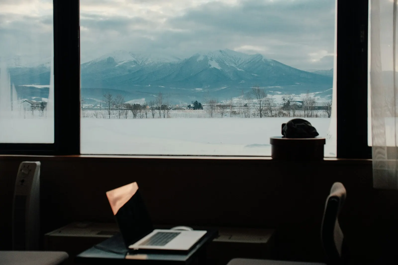 A laptop on a desk in front of a window looking out over snow-covered fields and mountains on a quiet winter workday