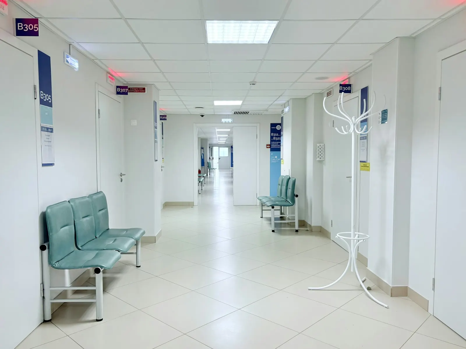 An empty, brightly lit hospital corridor with teal waiting chairs and numbered doors