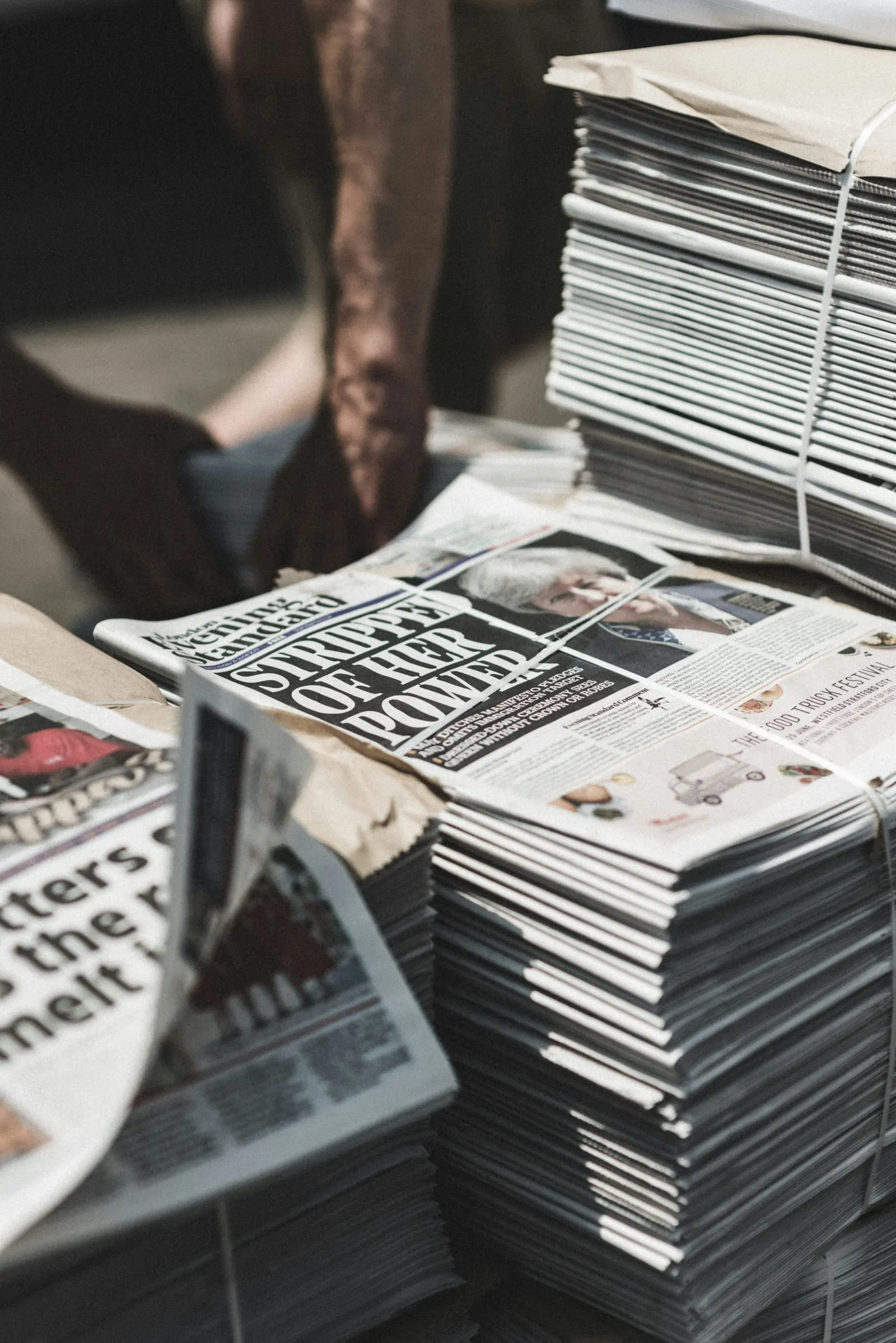 Bundled stacks of printed newspapers on a street stand, with a vendor's hands in the background
