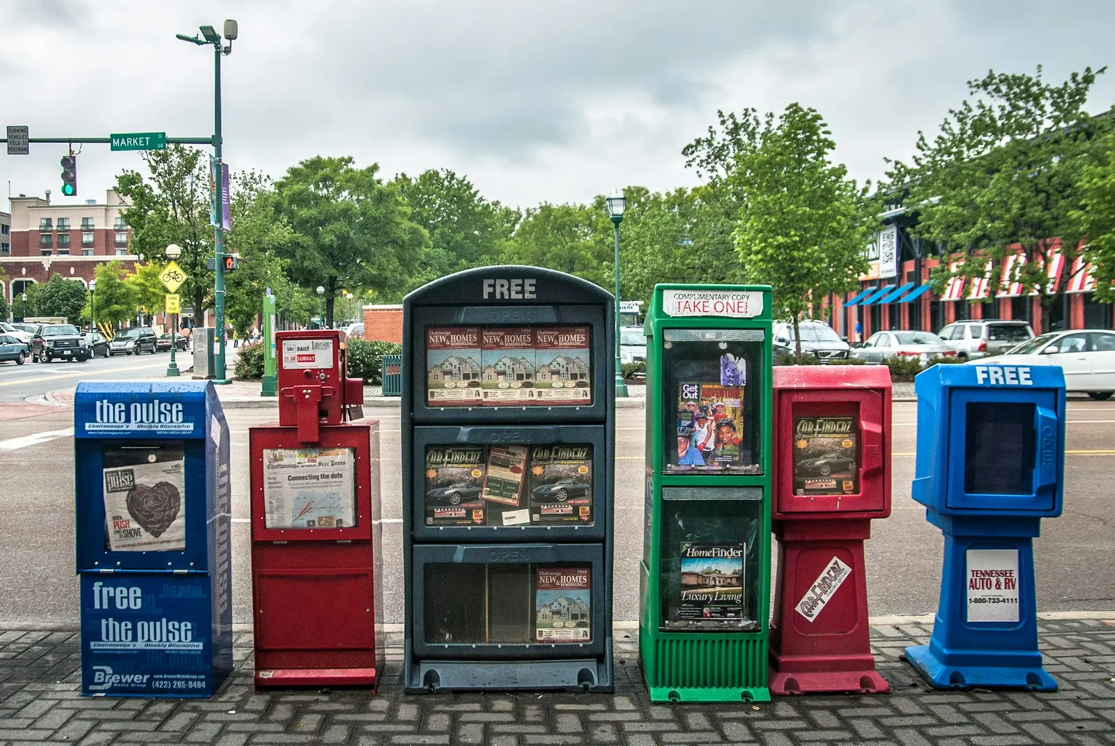 A row of free newspaper boxes on a city sidewalk, the regional media layer where many national stories begin