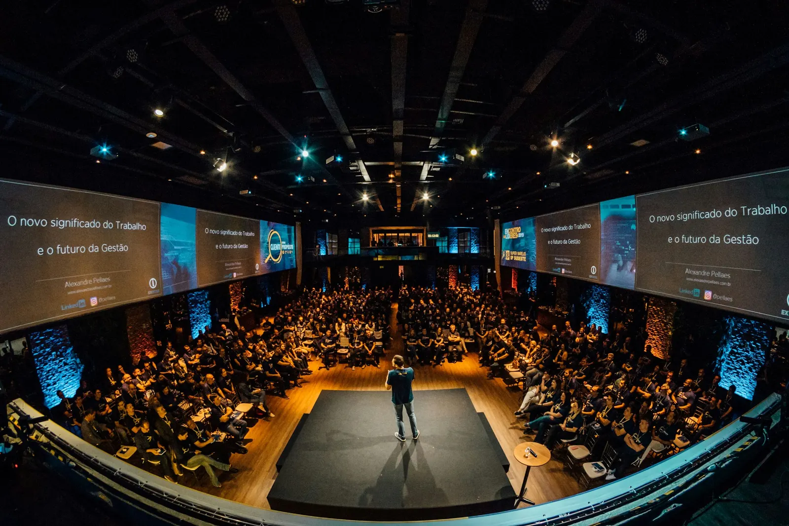 A speaker addressing a packed auditorium from a lit thrust stage