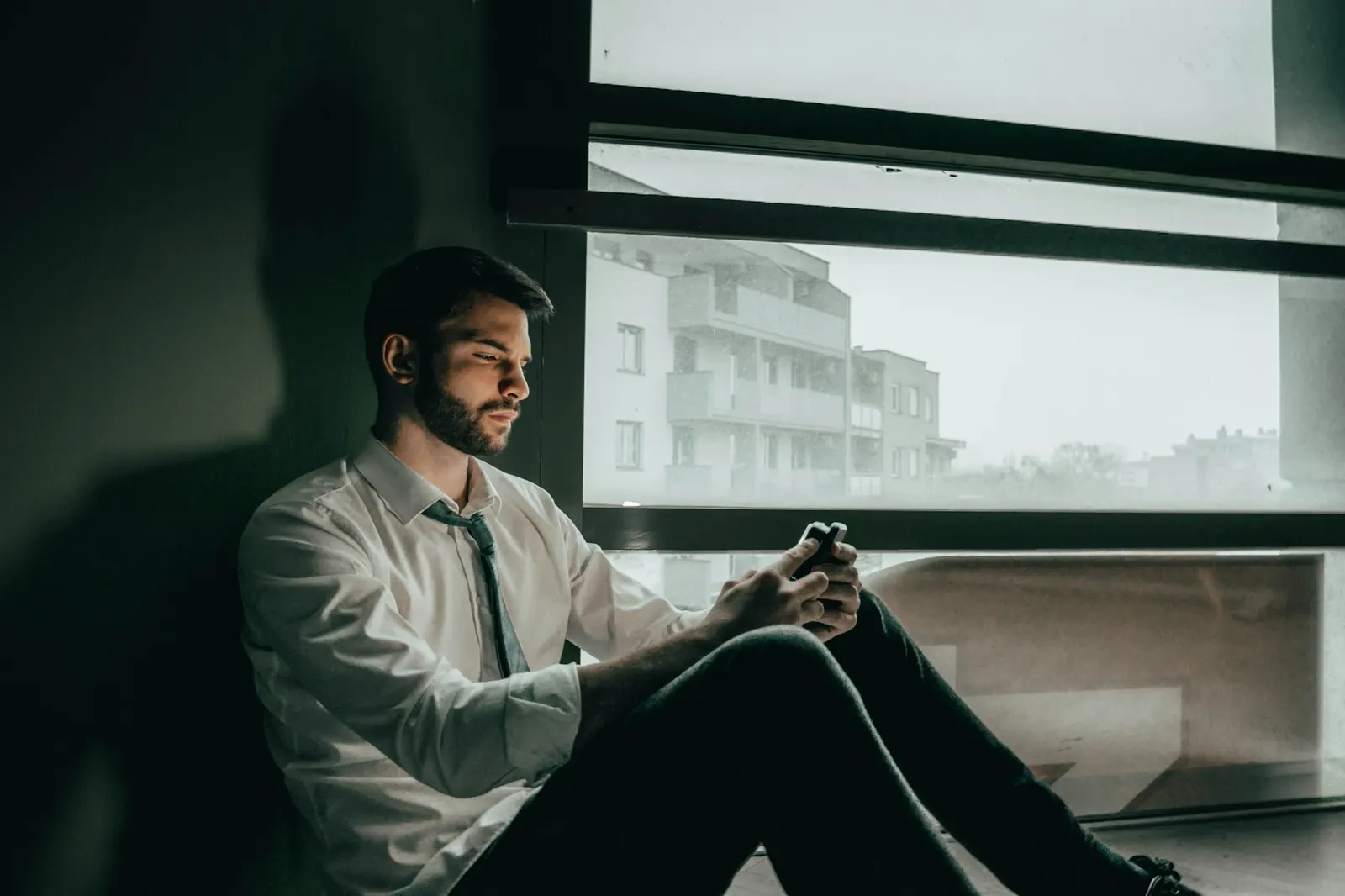 A professional in a shirt and tie reading on his phone beside a large window